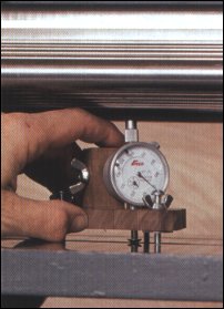 The planer bead gauge has to work in different positions. The base screw can straddle the bed slots. To check the feed rollers, orient the dial indicator, so it can take overhead reading. To check the bed rollers (shown here), flip the indicator on the tool post, sticking the plunger down through the hole in the base.